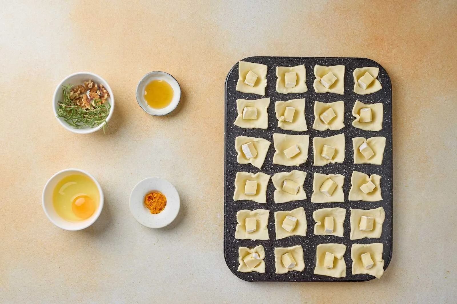 Brie cheese cubes placed inside puff pastry cups in a mini muffin pan while preparing Holiday Heaven Bites with rosemary, walnuts, honey, and orange zest.
