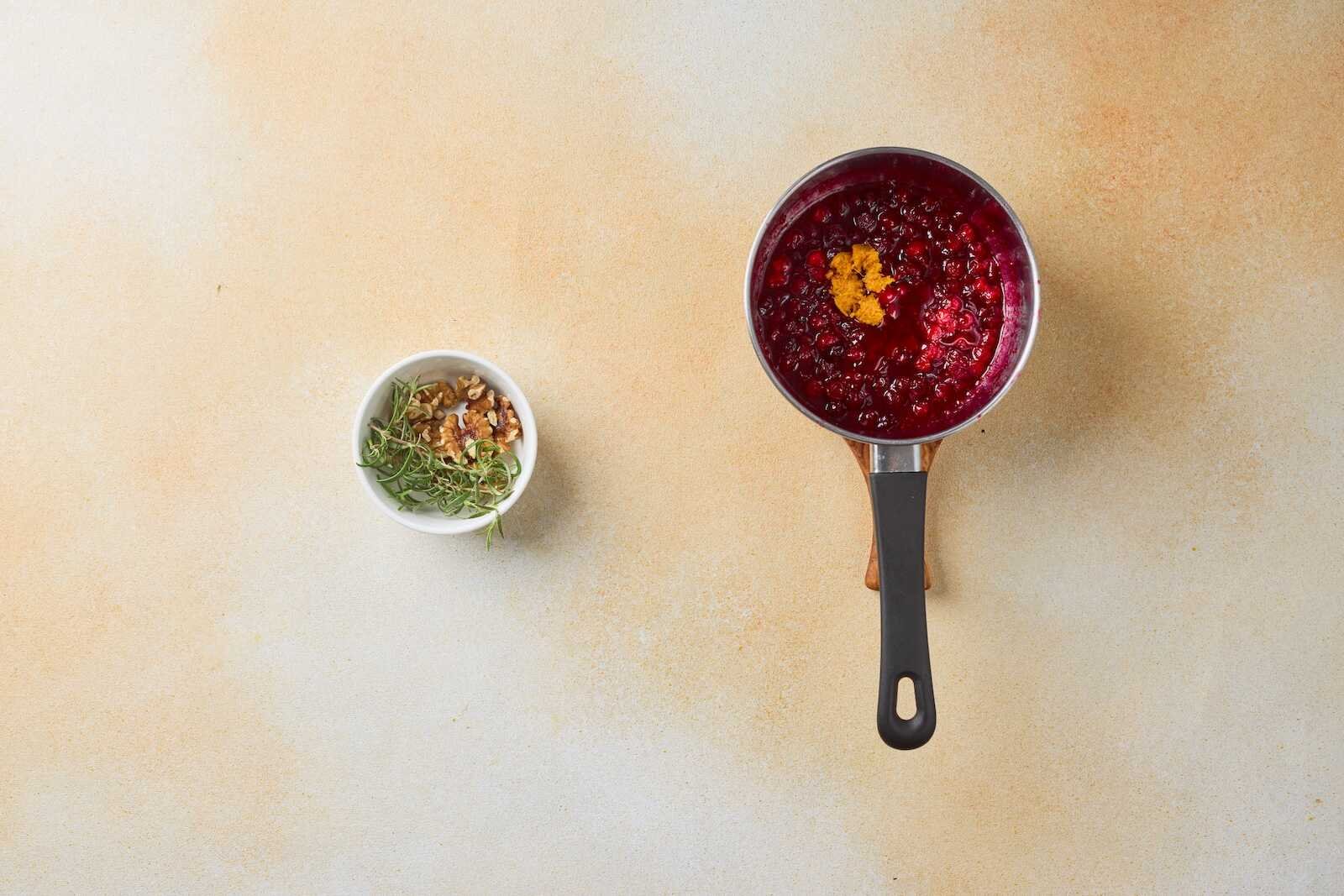 Homemade cranberry topping cooking in a saucepan with fresh orange zest for Holiday Heaven Bites, alongside chopped walnuts and fresh rosemary garnish.