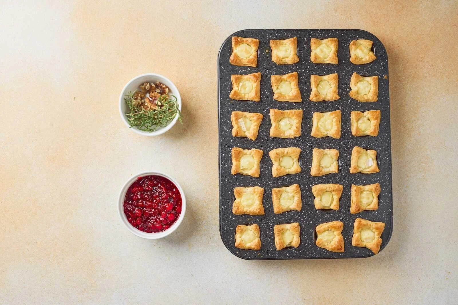 Freshly baked Holiday Heaven Bites with golden puff pastry and melted Brie cheese, ready to be topped with homemade cranberry sauce, walnuts, and rosemary.