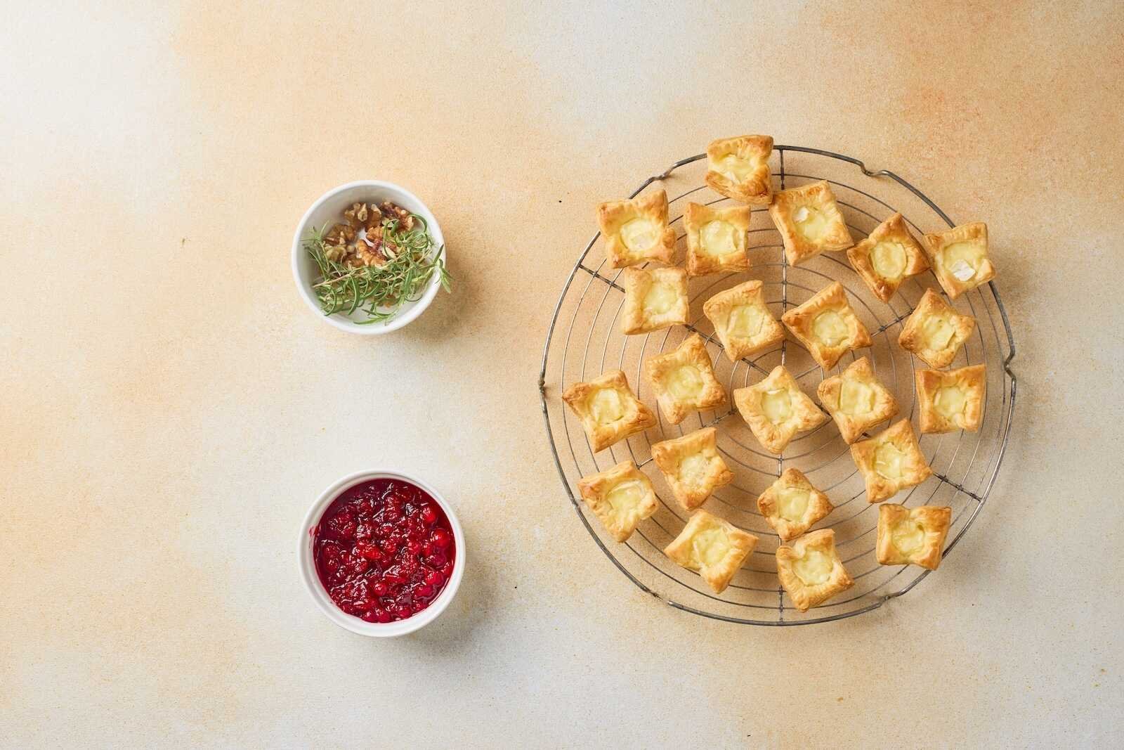 Baked Holiday Heaven Bites cooling on a wire rack with golden puff pastry, melted Brie cheese, homemade cranberry topping, walnuts, and fresh rosemary ready to assemble.