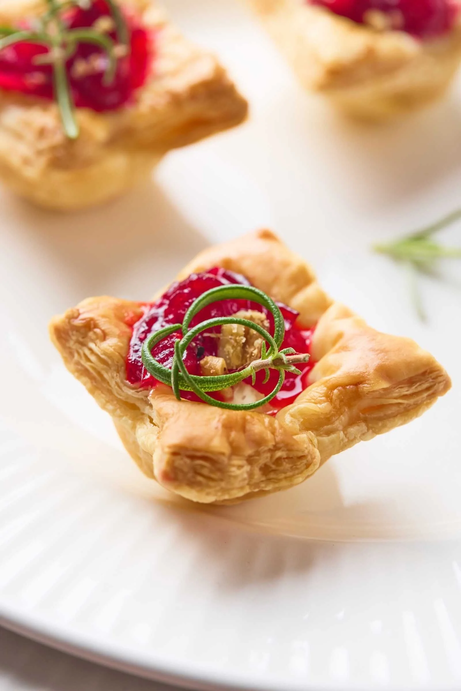 Close-up of a Holiday Heaven Bite with flaky puff pastry, creamy Brie cheese, homemade cranberry topping, chopped walnuts, and a fresh rosemary garnish.