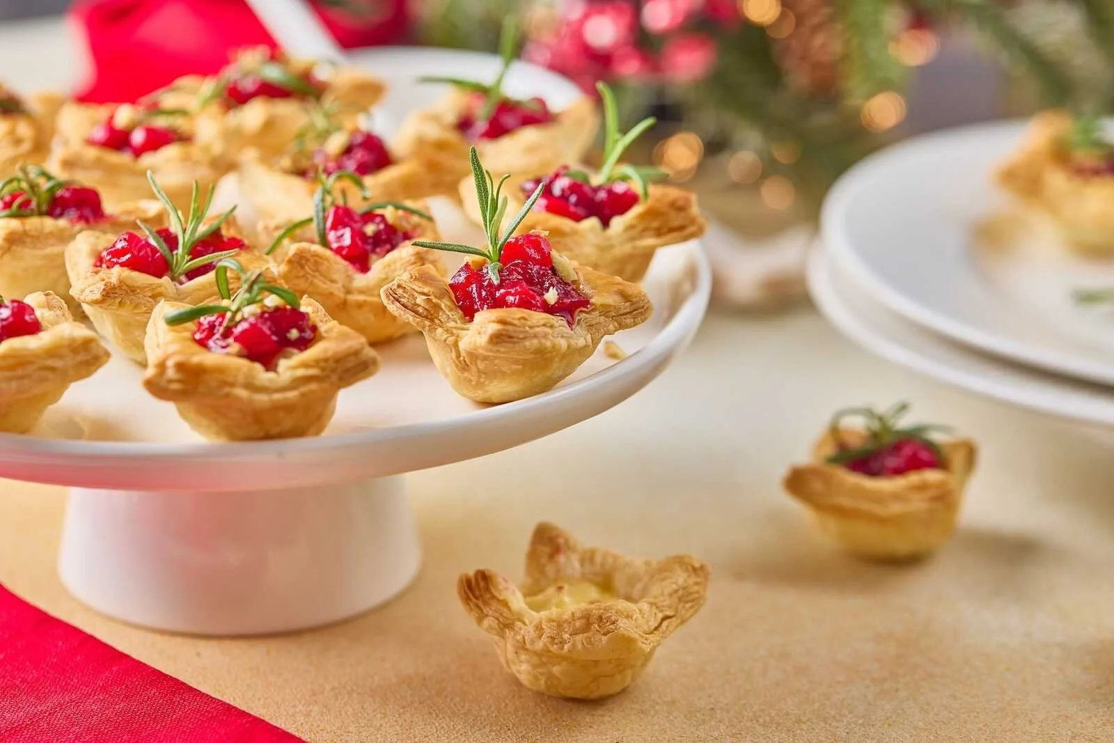 Holiday Heaven Bites displayed on a festive cake stand with flaky puff pastry, creamy Brie cheese, homemade cranberry topping, chopped walnuts, and fresh rosemary garnish.