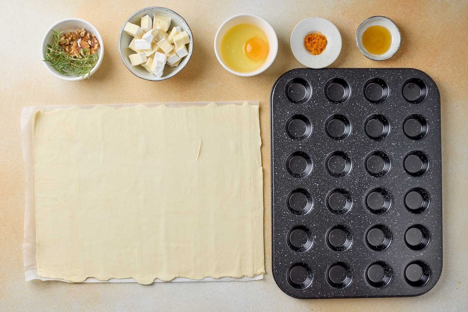 Ingredients and equipment for Holiday Heaven Bites including puff pastry, Brie cheese, walnuts, rosemary, egg, orange zest, honey, and a mini muffin pan.