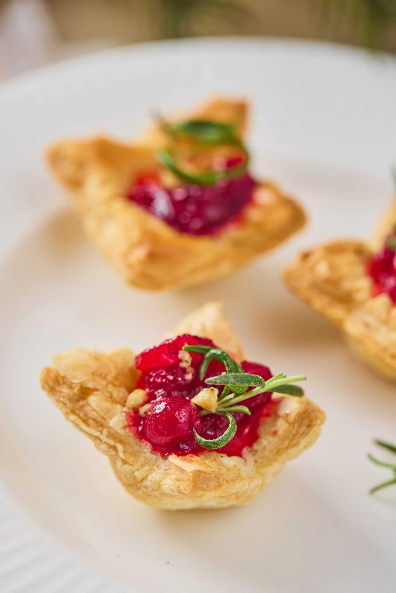 Close-up of a Holiday Heaven Bite with flaky puff pastry, creamy Brie cheese, homemade cranberry topping, chopped walnuts, and fresh rosemary on a white serving plate.
