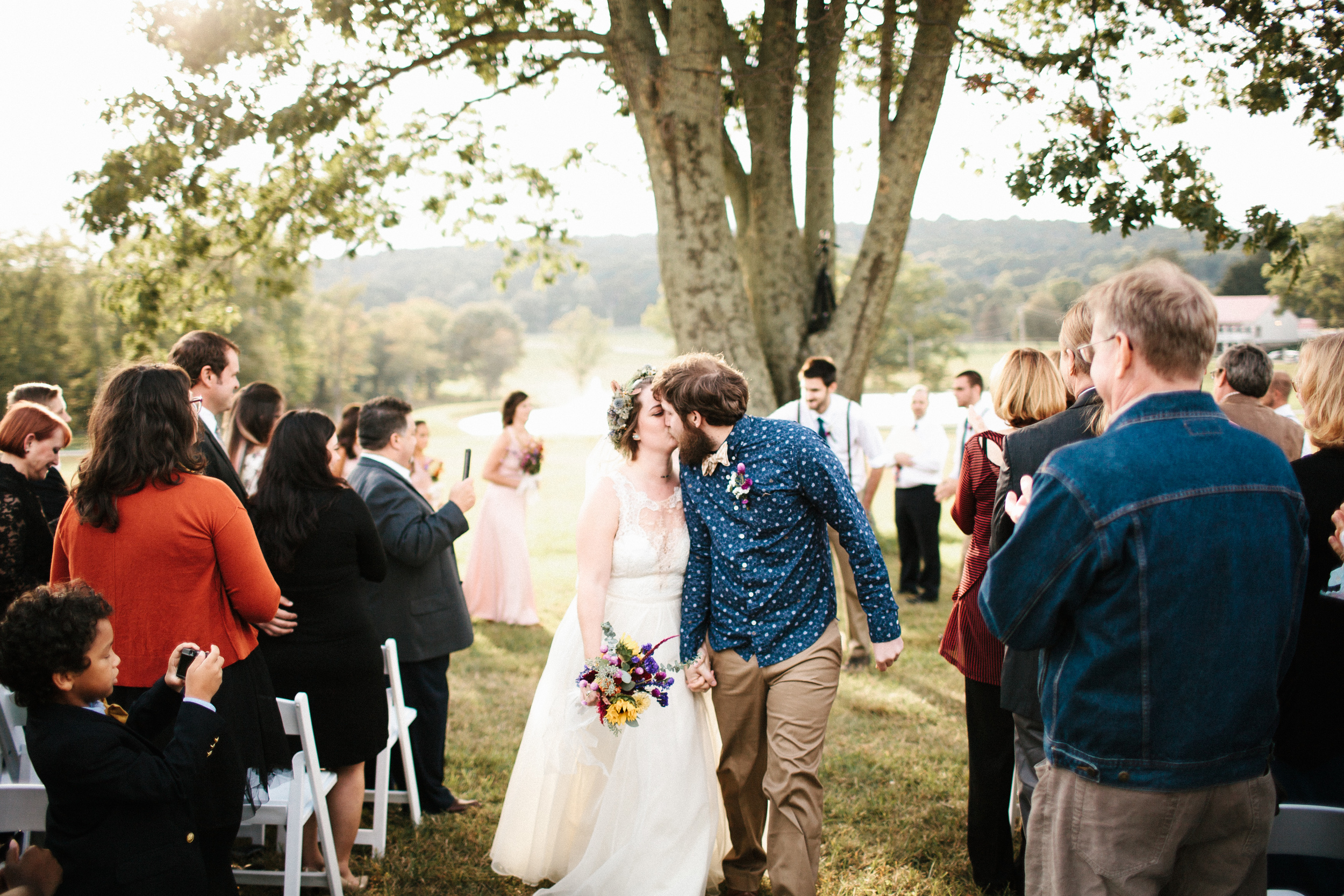  Bride and groom kiss in front of tree in wedding aisle. 