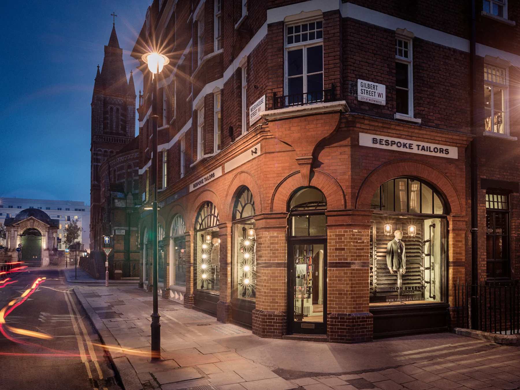 Street corner with brick building housing a boutique clothing store named 'Bespoke Tailors', window mannequins dressed in suits, at dusk in Westminster, London, with street signs for Gilbert Street and Weighthouse Street.
