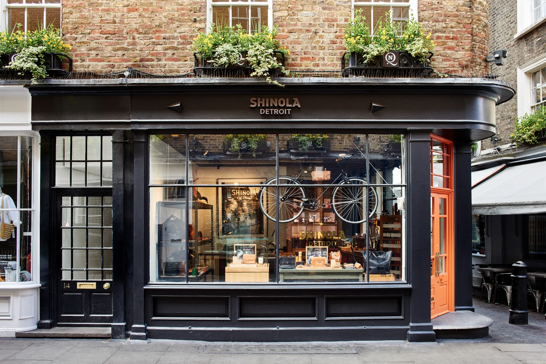 A storefront named Shinola Detroit featuring large glass windows and a brick building facade. Inside, a bicycle is displayed hanging above the window, and various accessories and products are visible. The exterior has a black and orange color scheme,
