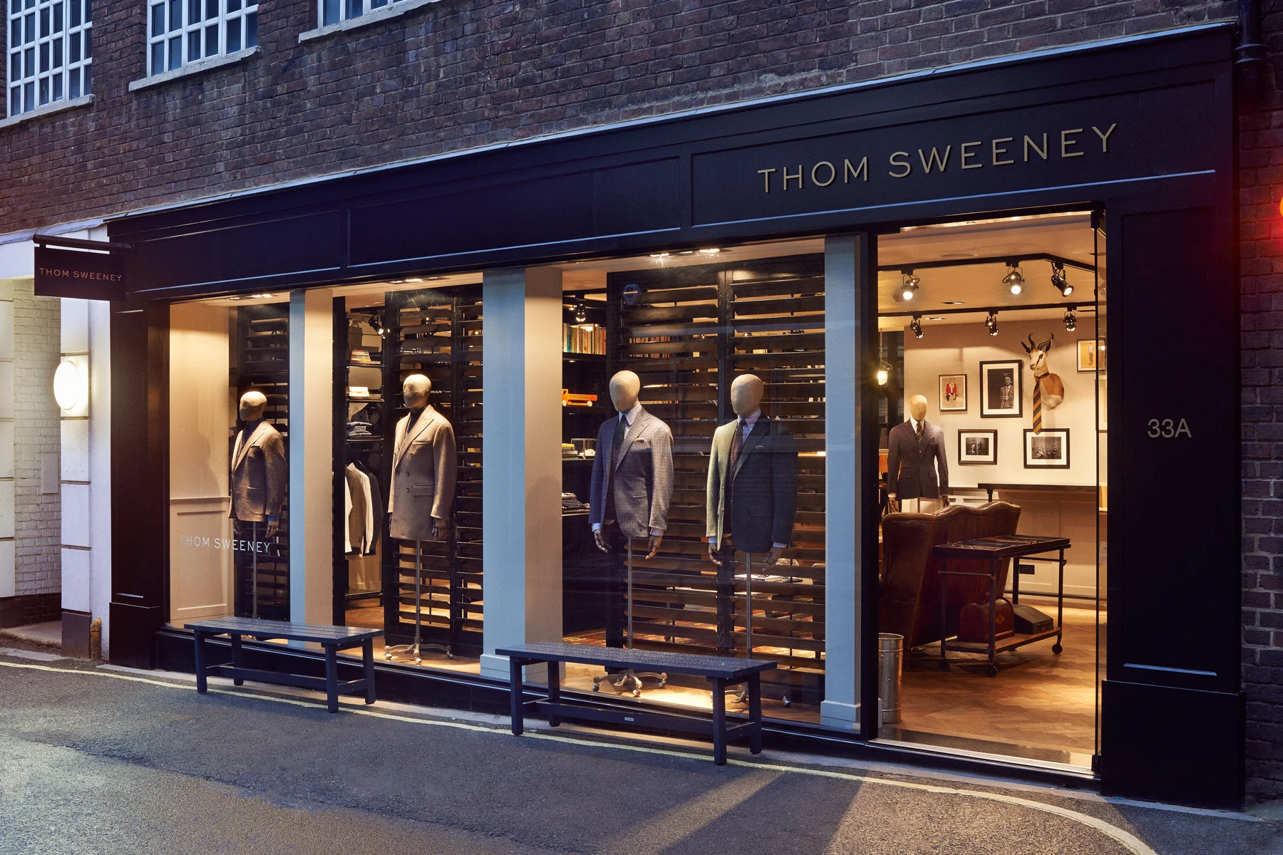 Storefront window display of Thom Sweeney featuring mannequins dressed in suits, with bookshelves and framed pictures inside, and mounted deer head on the back wall.