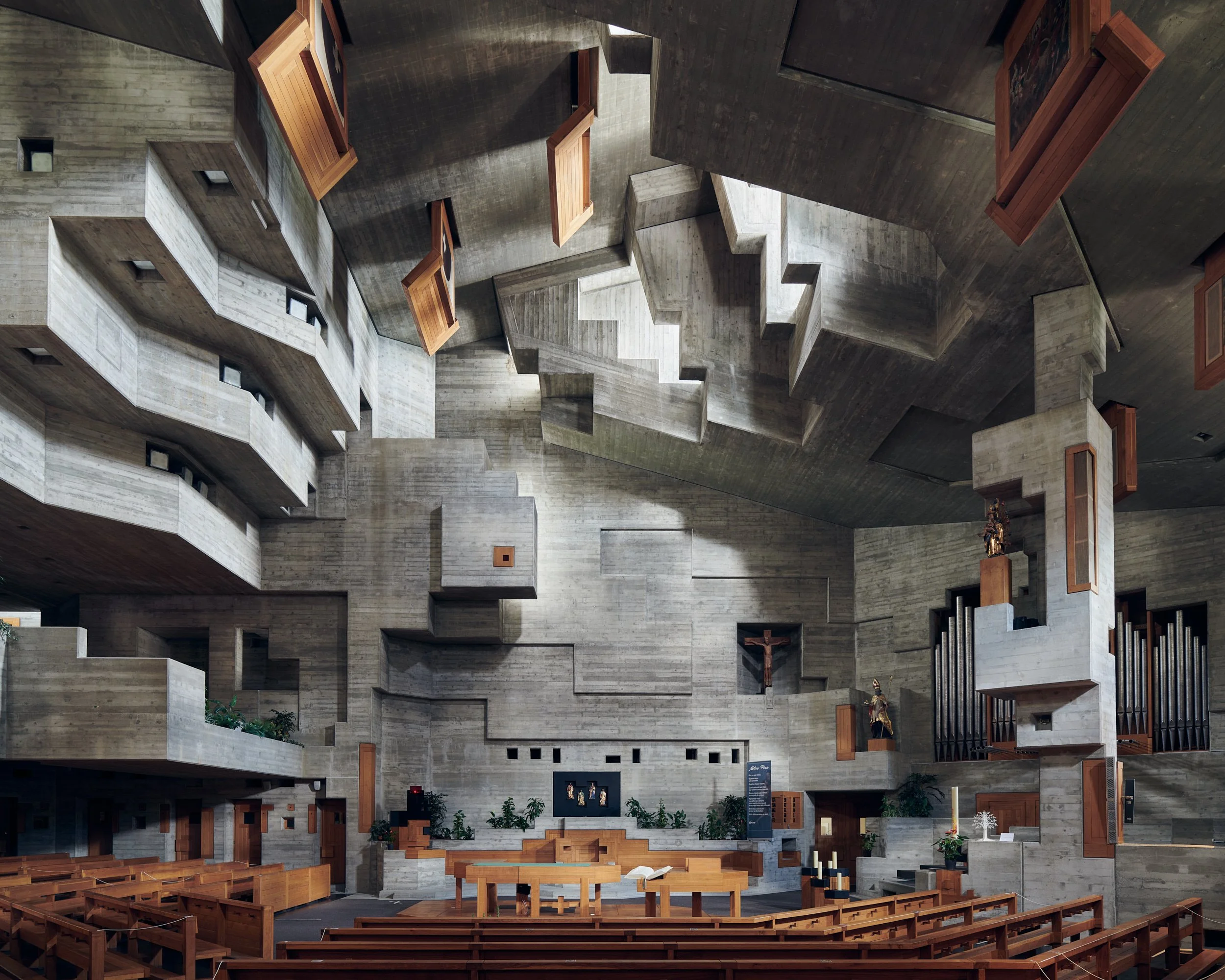 The interior of a modern church with concrete walls, wooden pews, a large cross, candles, and religious artwork.