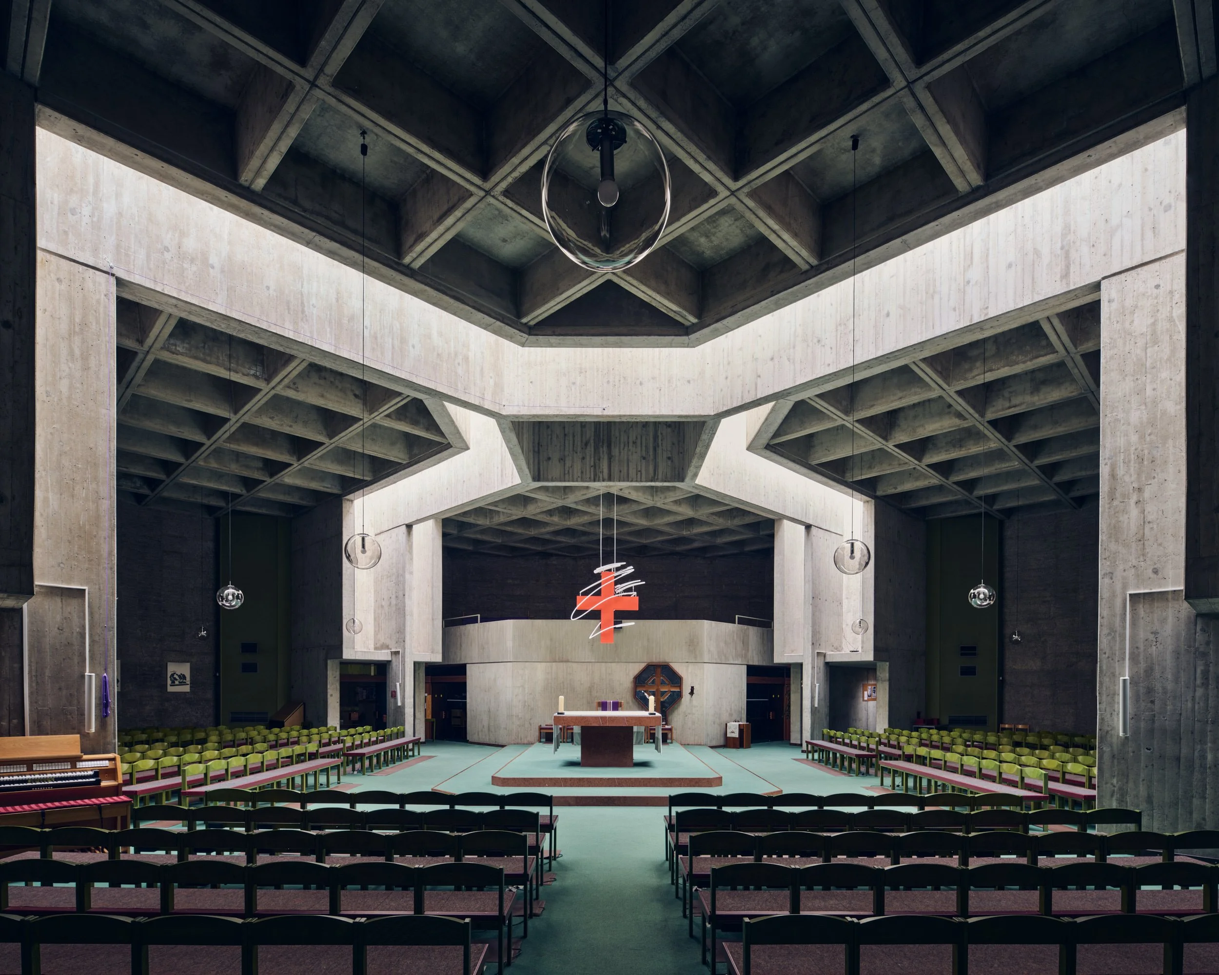 Interior view of a church or cathedral with concrete architecture, rows of green chairs, and a stage with a cross at the front. Modern lighting fixtures hang from the ceiling.