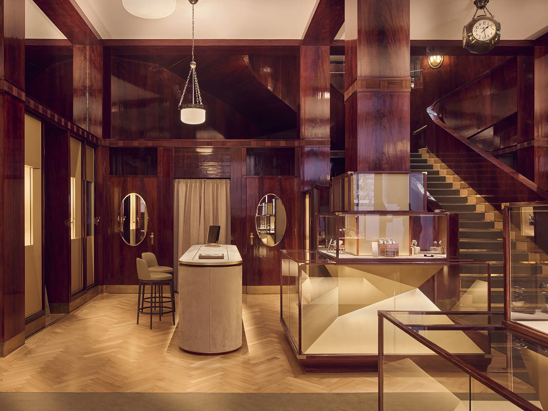 Interior of a luxurious jewelry store with polished wood walls, display cases with jewelry, a staircase, and a seating area with chairs and a table.