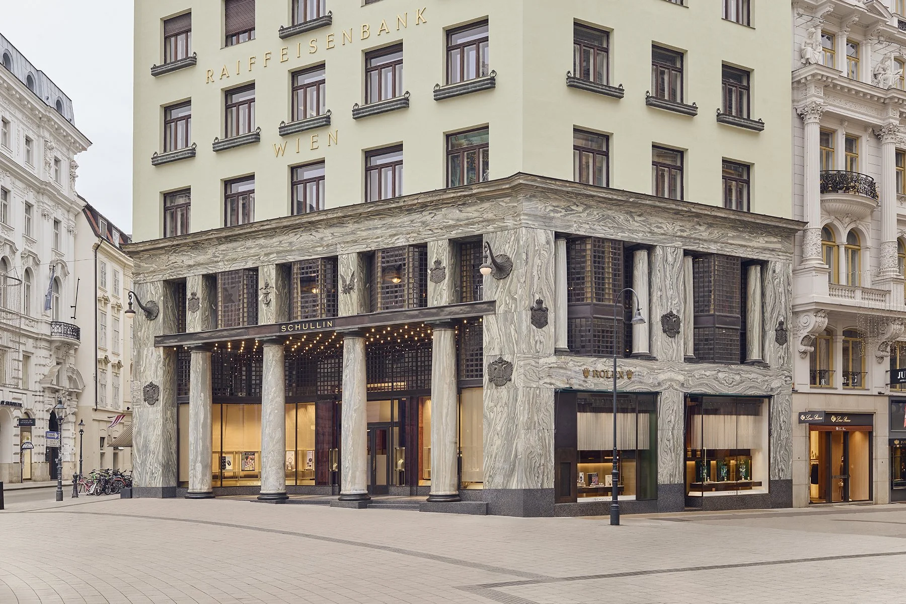 A modern building with a marble facade and columns on a city street, with signs for Rolex and Schullin, and surrounding historic white buildings.