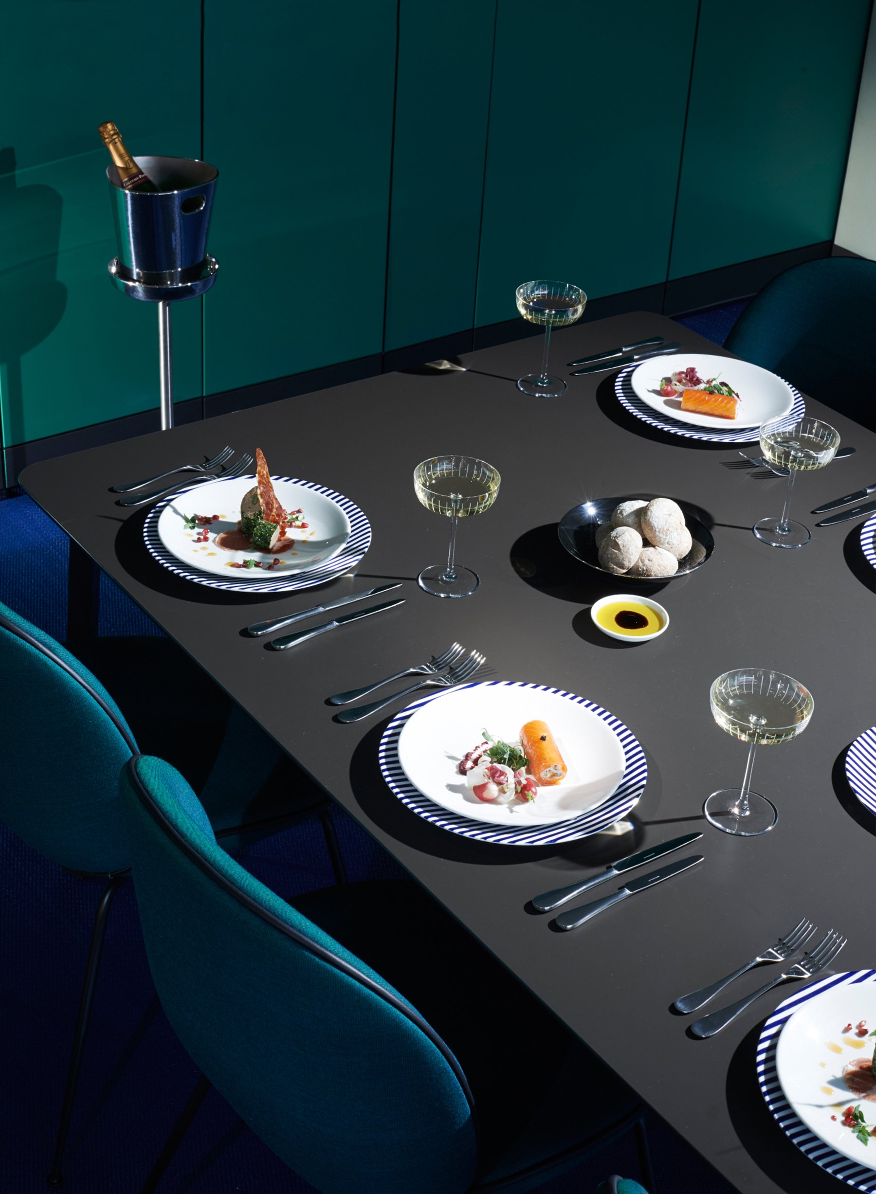 A dining table set with plates of gourmet food, wine glasses, and silverware, with teal chairs surrounding it and a teal cabinet in the background.