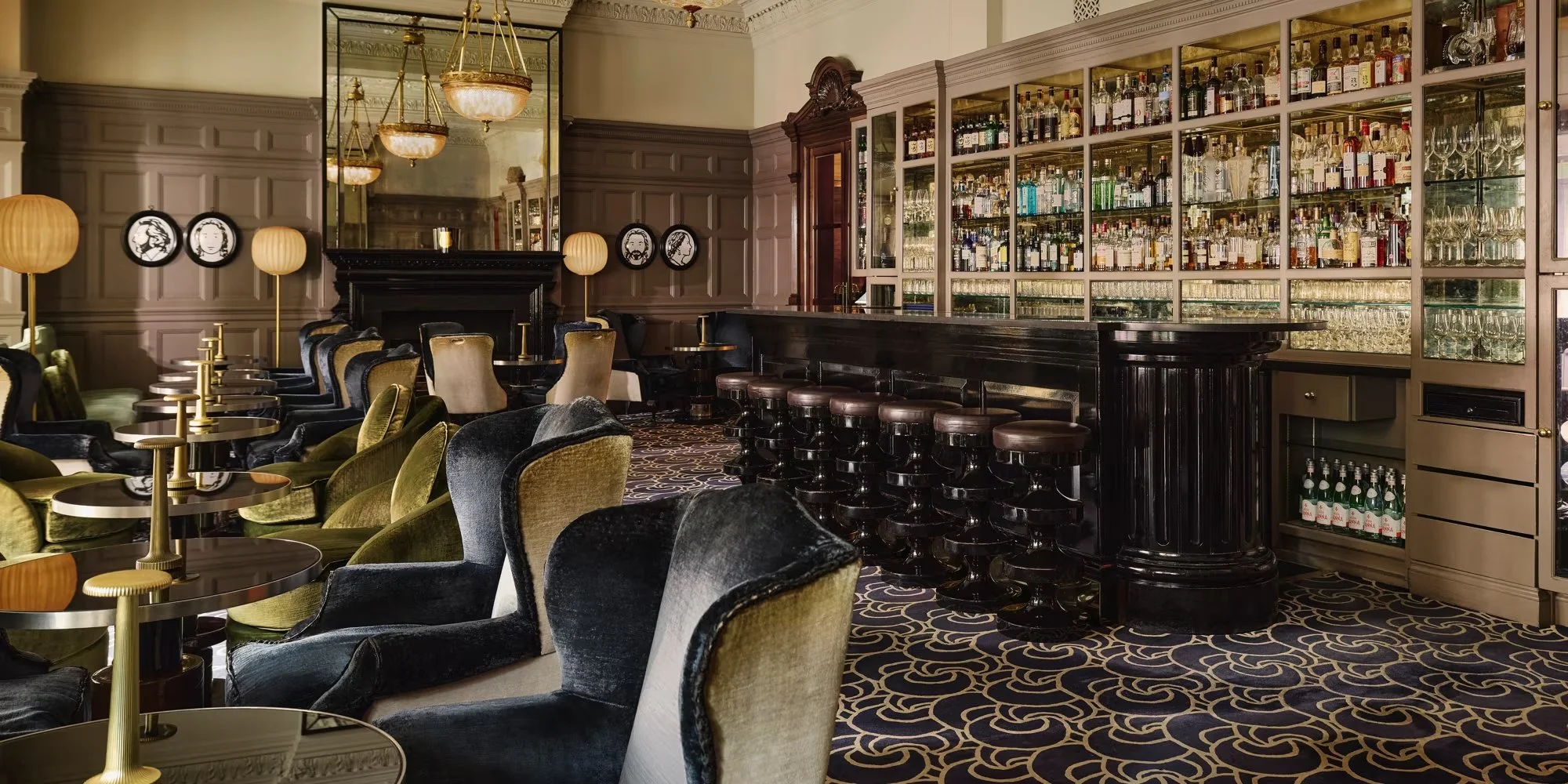 A luxurious bar and lounge area with plush velvet chairs, gold lamps, a dark wooden bar counter, and glass shelves filled with various bottles of alcohol.