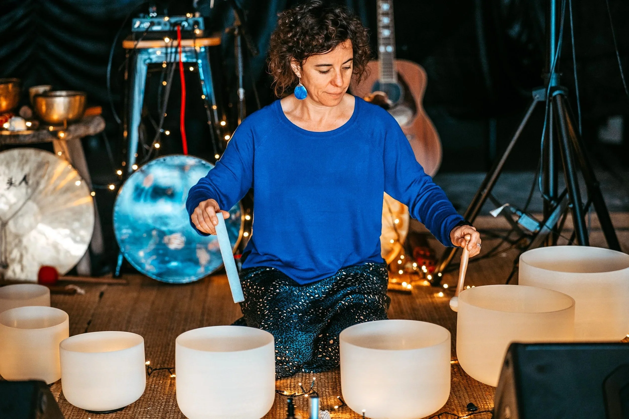 A woman dressed in a blue top and patterned pants playing crystal singing bowls surrounded by musical instruments and fairy lights.
