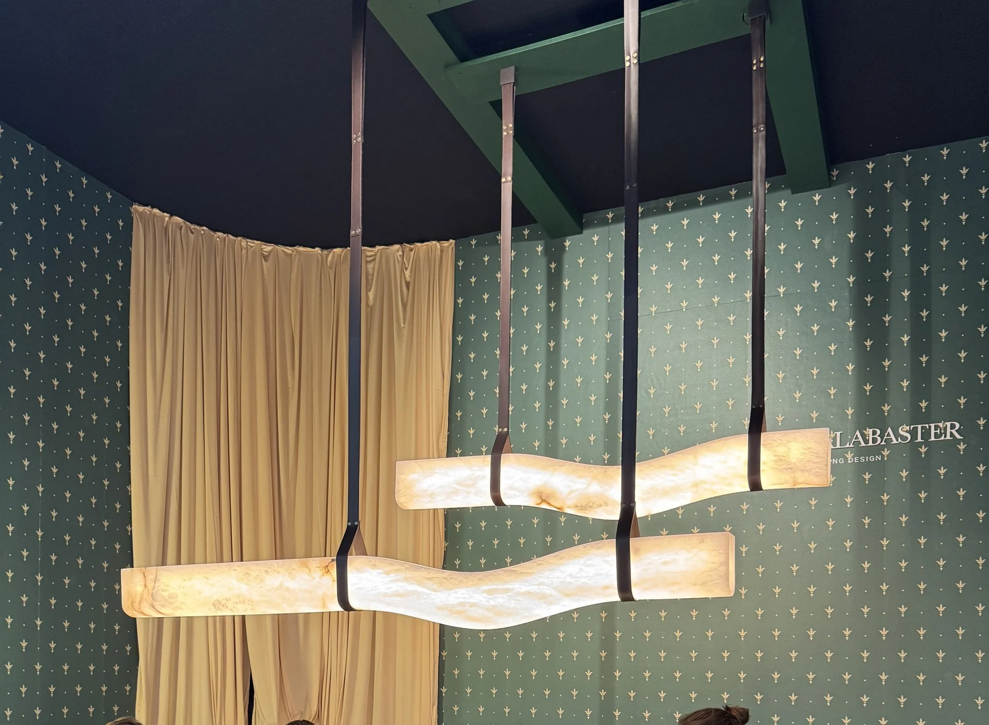Two long horizontal alabaster pendants glowing above an oval dining table on a green stand, United Alabaster at Decorex 2026