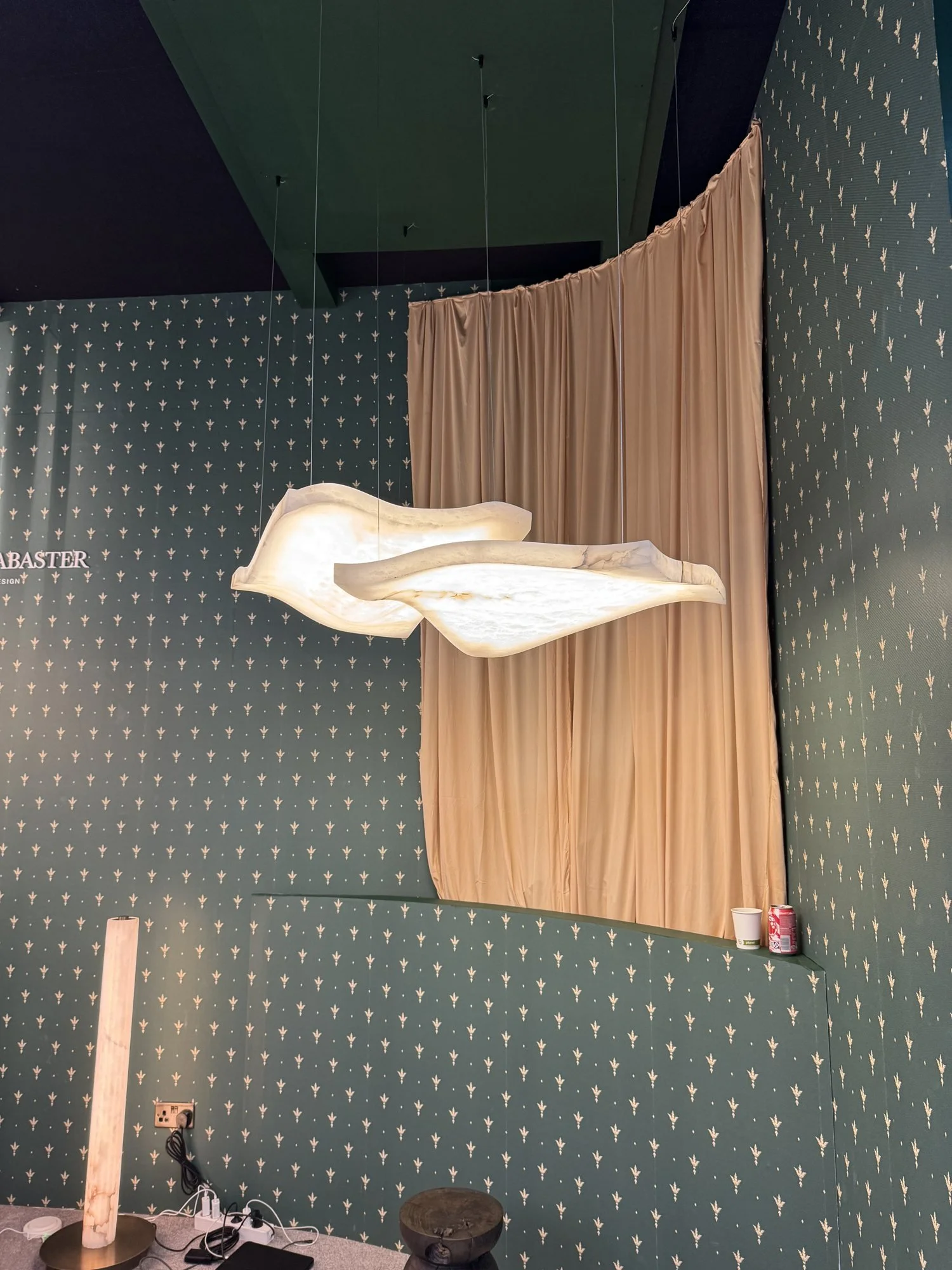 A wave shaped alabaster pendant hanging in front of a peach curtain on a green patterned wall, United Alabaster at Decorex 2026
