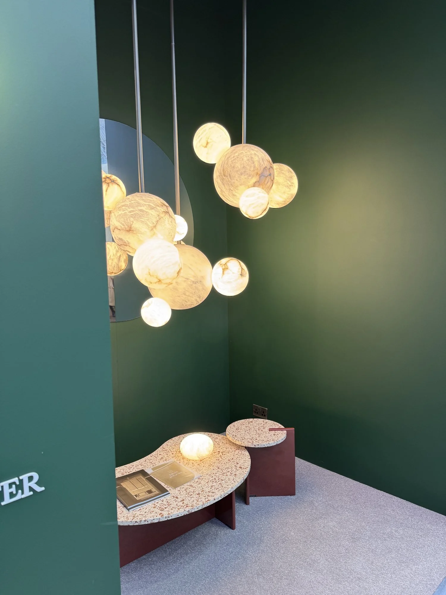 A cluster of round alabaster globe pendants lit against a dark green wall, United Alabaster at Decorex 2026