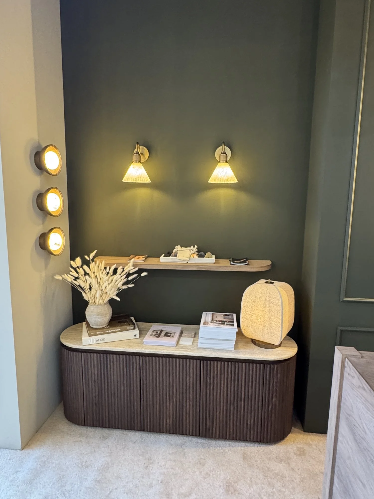 A fluted dark wood sideboard in an olive green alcove, with glass wall lights and round wall lights above, Industville at Decorex 2026