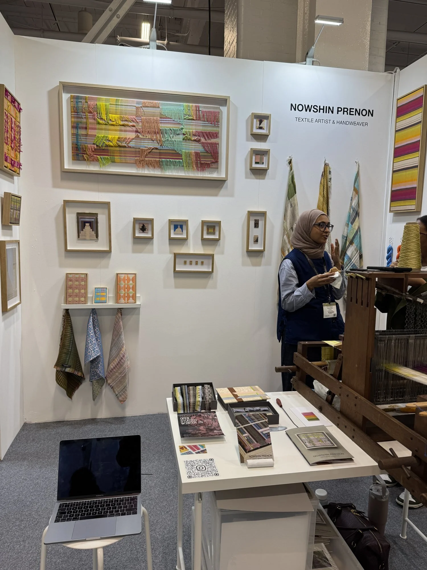 A white stand with framed handwoven pieces, hanging textiles and a loom, with the weaver talking to a visitor, Nowshin Prenon in Making Spaces at Decorex 2026