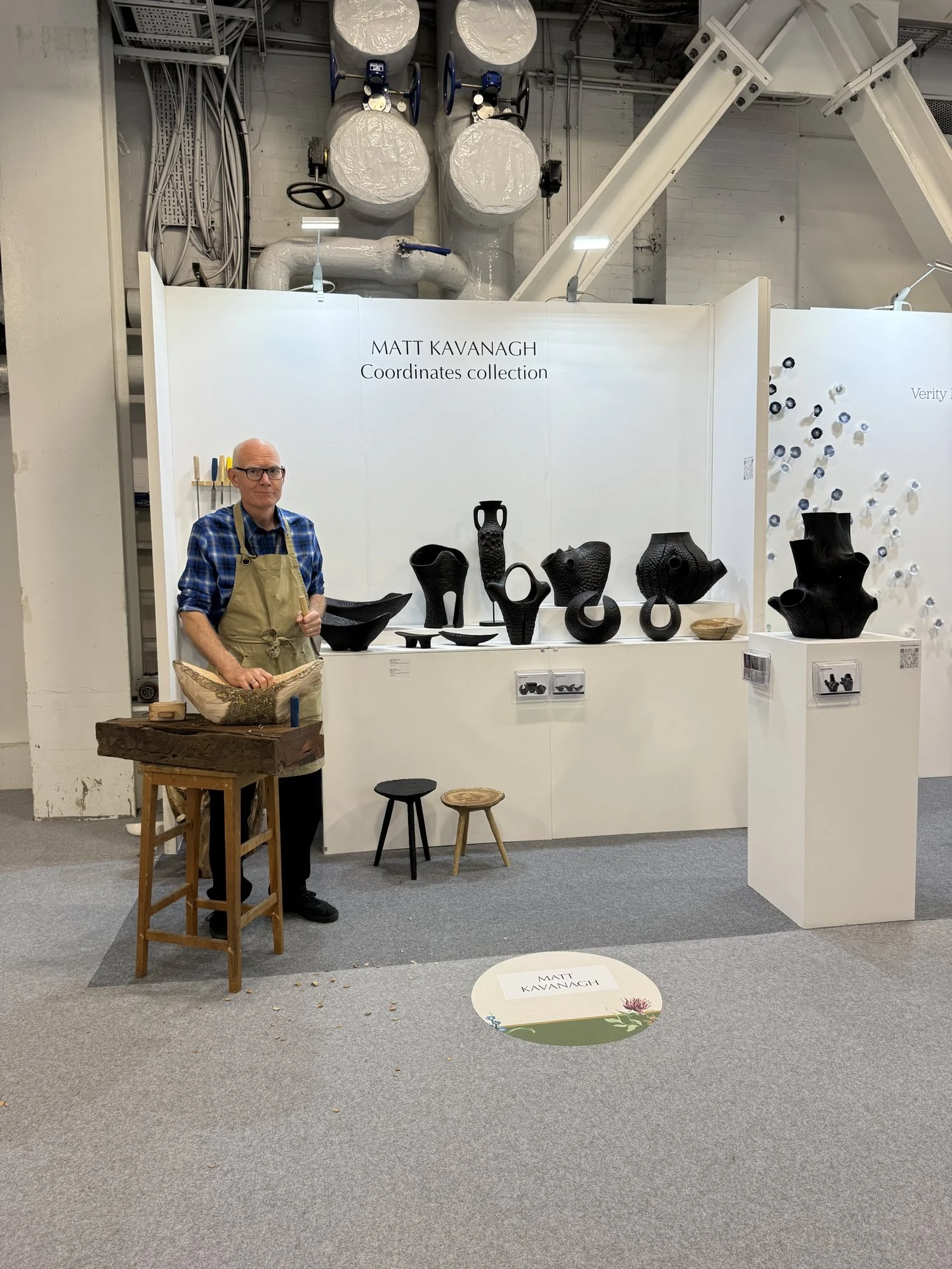 Dark carved wooden vessels on a white plinth, with the maker at his workbench, Matt Kavanagh in Making Spaces at Decorex 2026