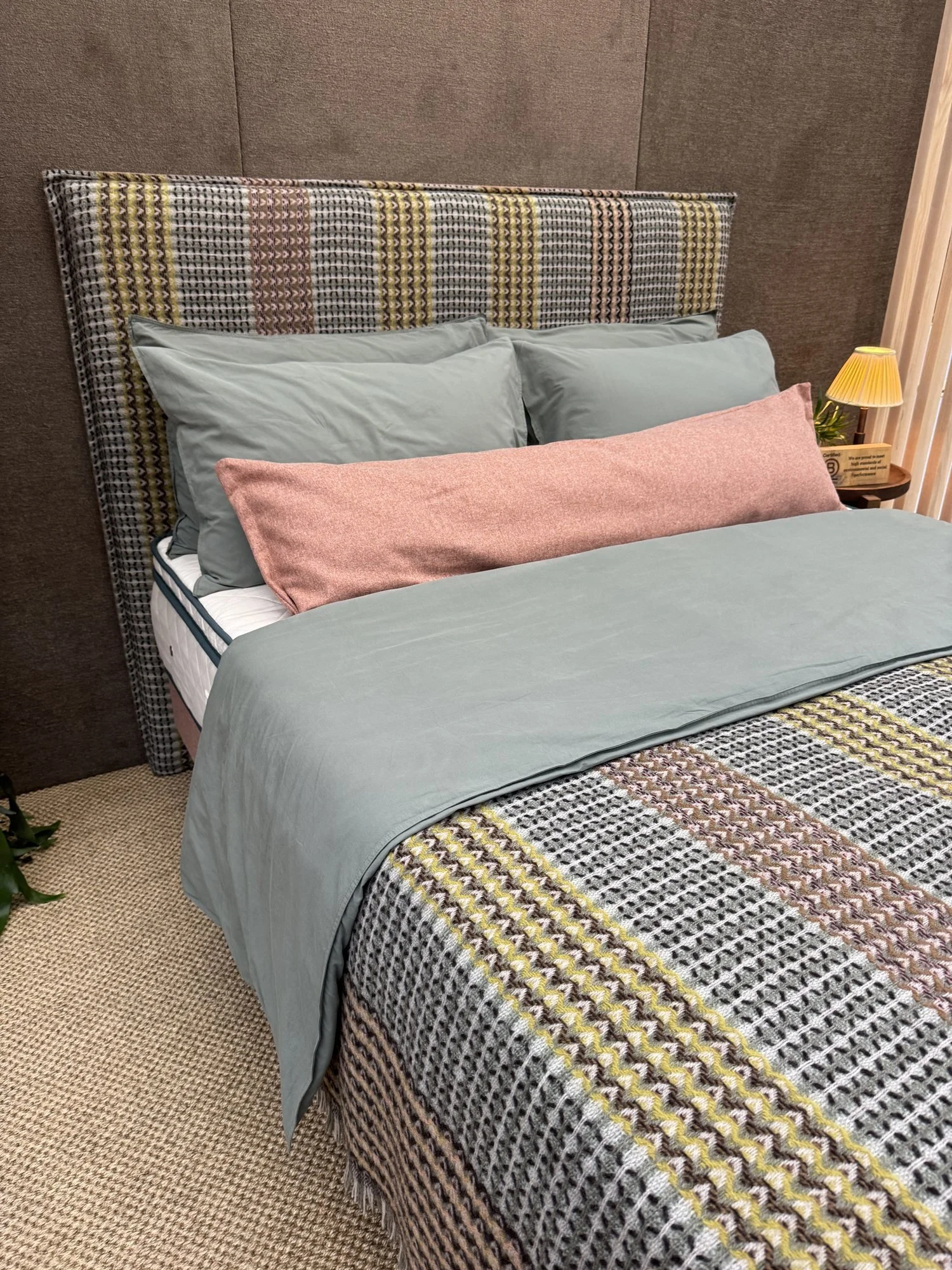 A bed with a tall woven check headboard in yellow, pink and grey, sage bedding and a pink bolster cushion, Naturalmat at Decorex 2026