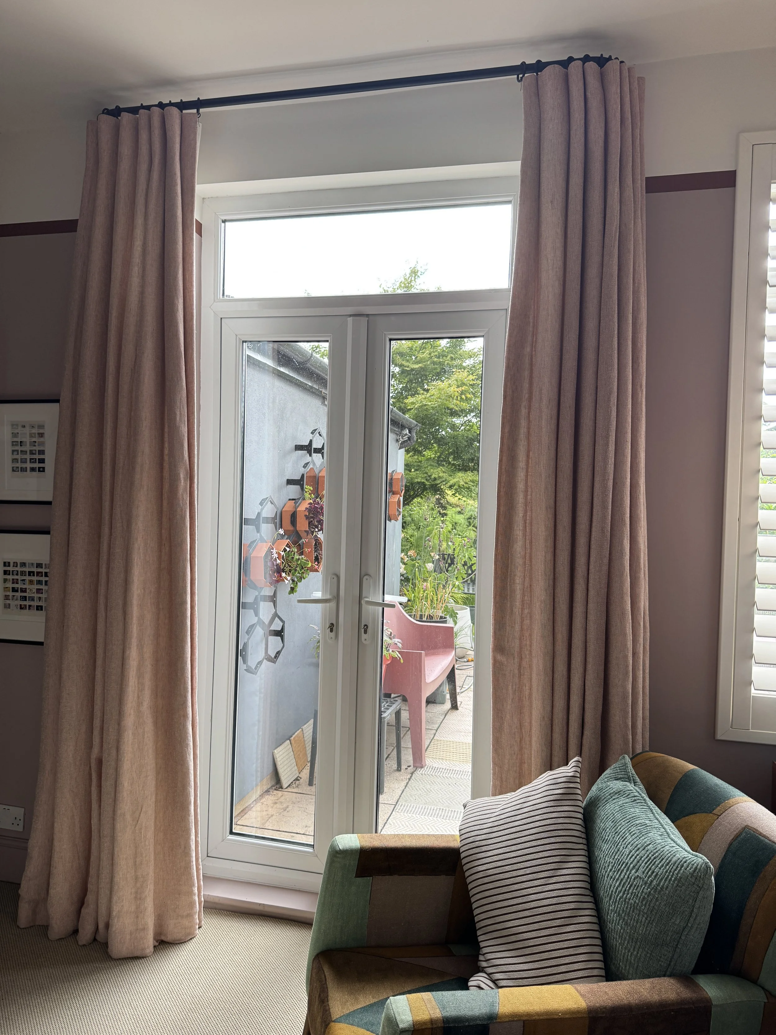 Bespoke Bouclin curtains by Kvadrat in a soft blush tone, hung on a black French pole above glazed doors in a dining room and interior design studio. The curtains are designed to blend seamlessly with the wall colour and maximise natural light.