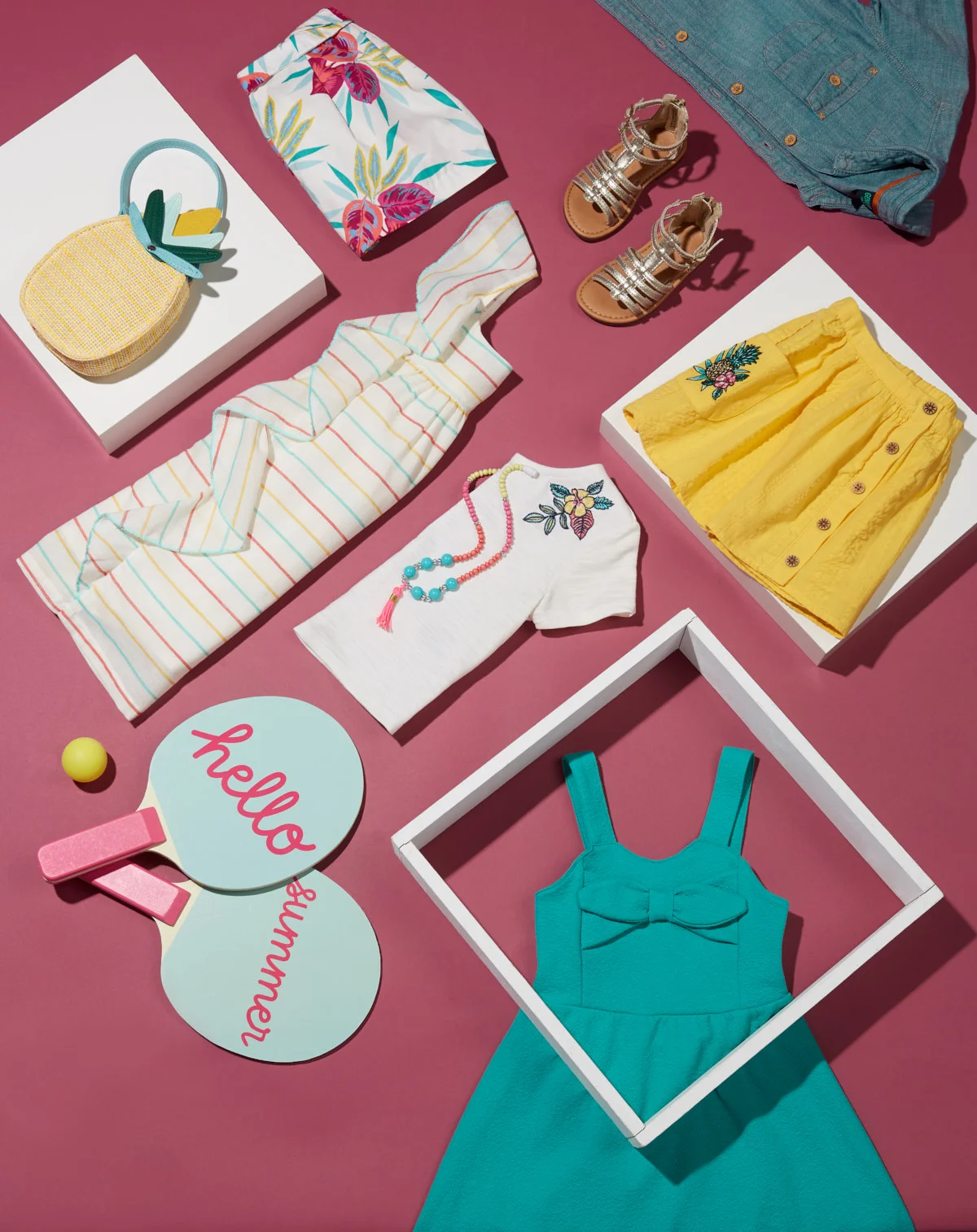  Studio photo of a summer collection of girls kids clothing and accessories in blues and yellows laying on a rasberry background. Apparel is arranged at a diagonal to create a lively graphic look. Photography: Leah Perry, Styling: Cyn Ng 