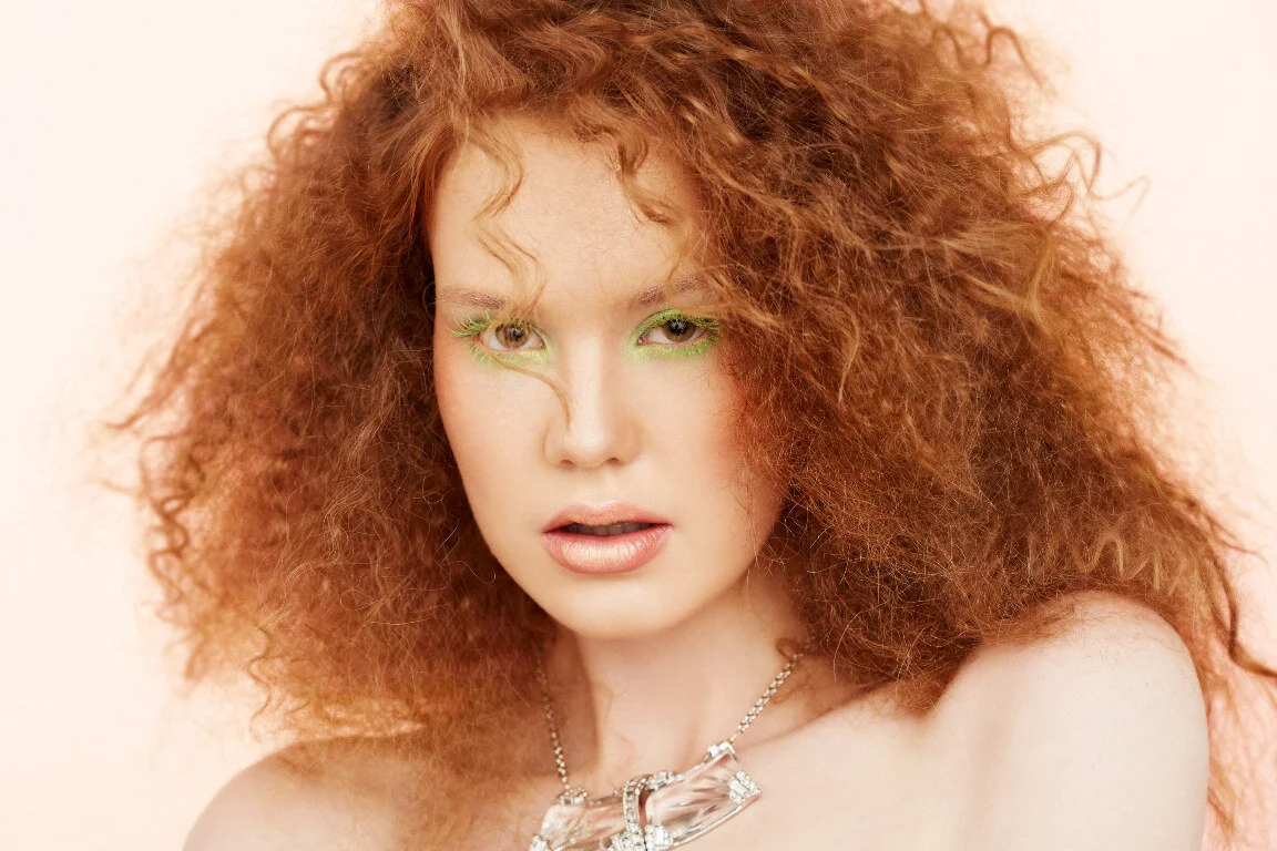  Pastel themed beauty shoot. Model with big messy curly red hair shot on pale pink, with frosted lipstick and lime green rimed eyes. She wears a statement necklace of clear acrylic pieces.  Photography: Leah Perry Photography  Hair and Makeup: Saj Ma