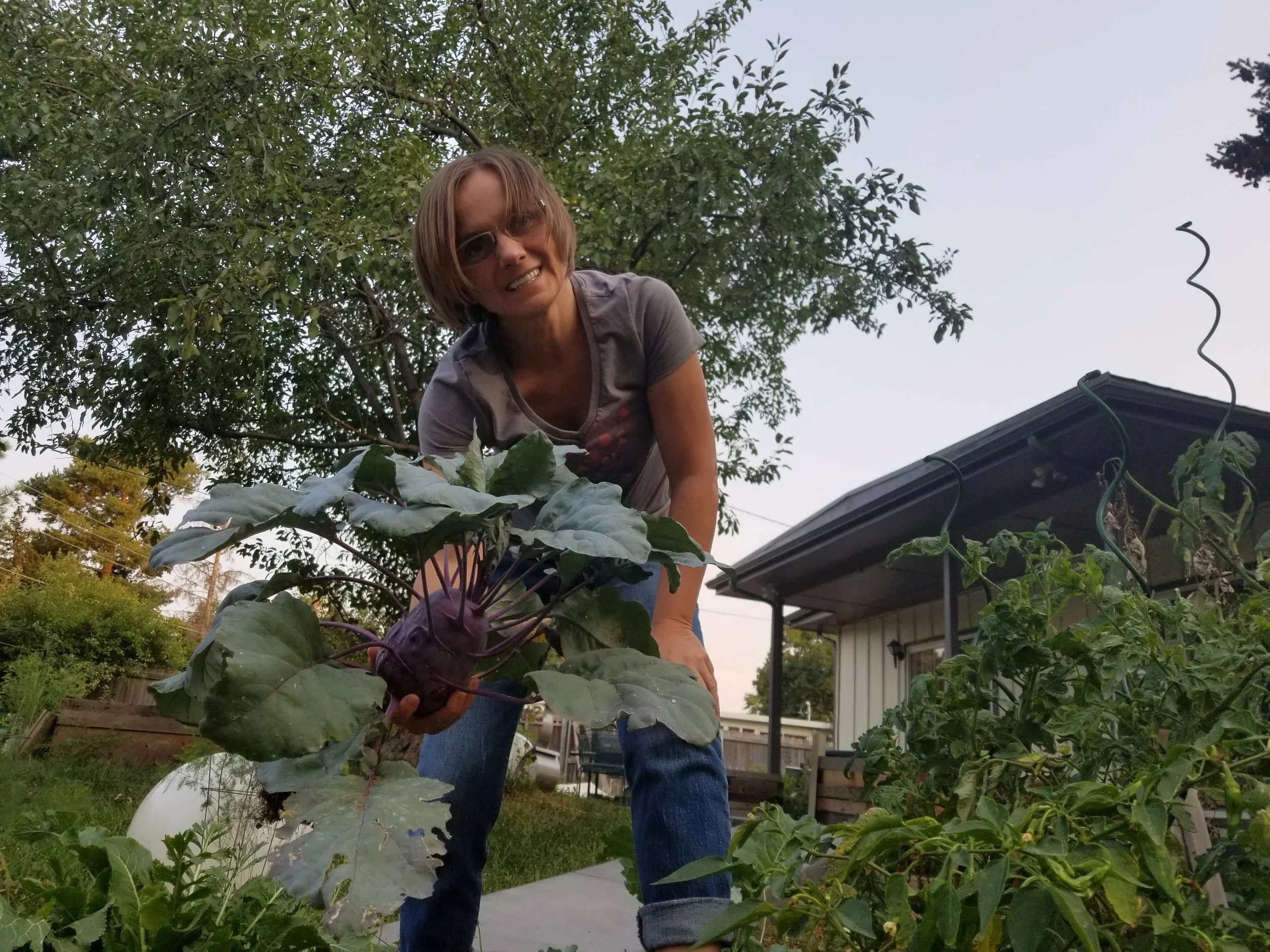 A smiling woman with glasses, holding a large purple kohlrabi, in a garden with various plants, trees, a house, and a cloudy sky in the background.