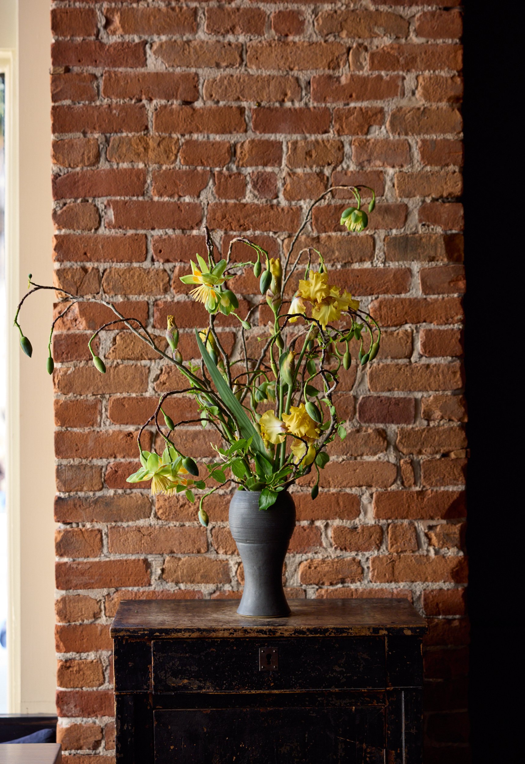 Yellow flowers in a Vase placed on a dining room hutch next to a red brick wall.