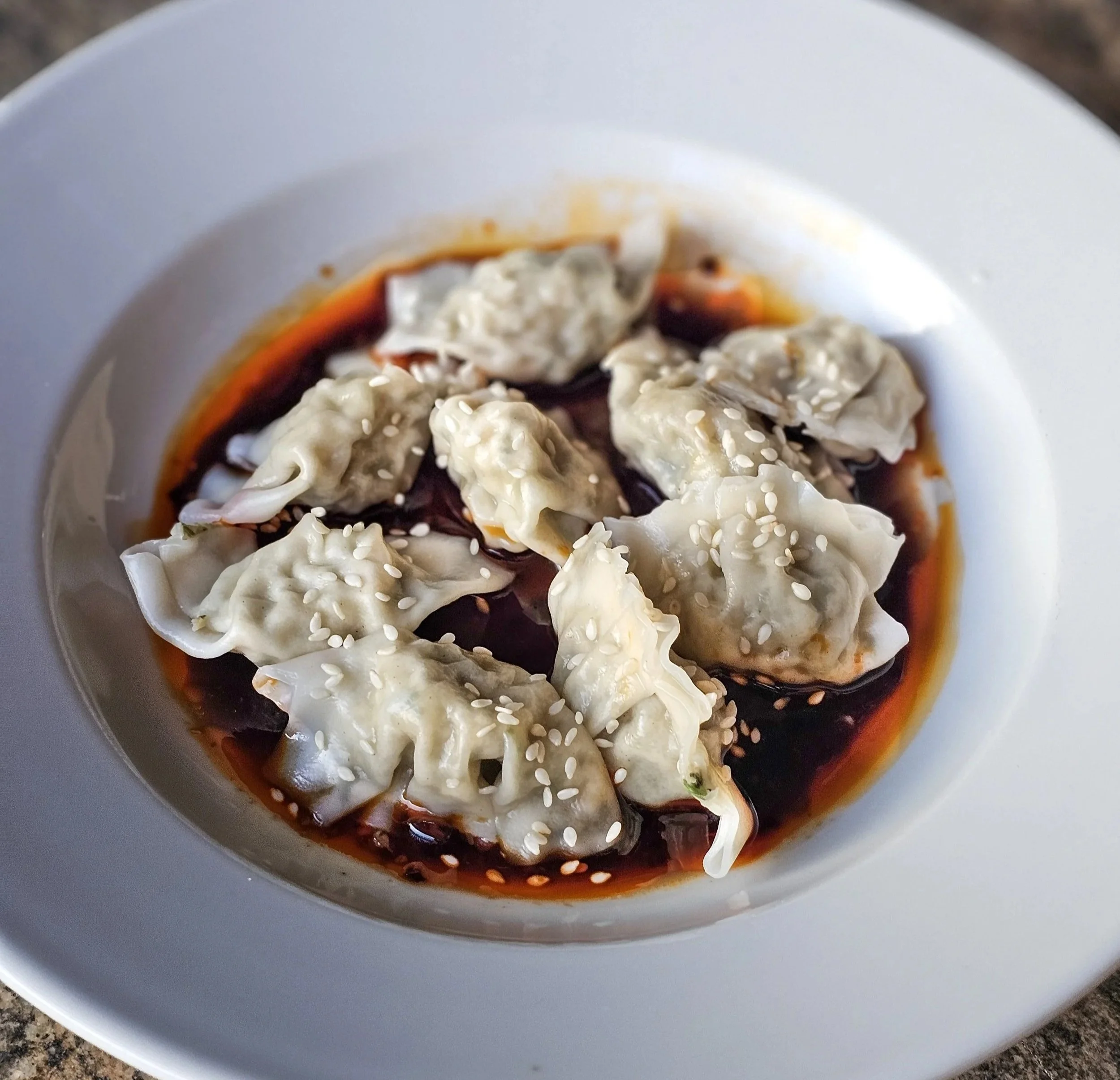 Sichuan-style dumplings served in chili oil, topped with sesame seeds and arranged in a shallow white bowl.
