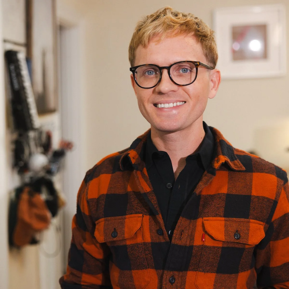 A smiling man with blonde hair and glasses wearing a black shirt and orange plaid jacket, standing indoors near a wall with framed art.
