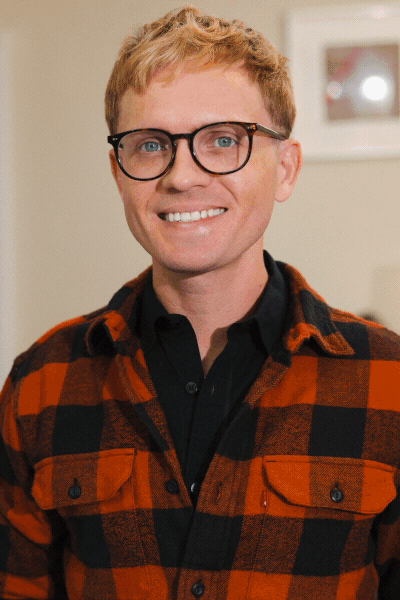 A smiling man with red hair, glasses, wearing a black shirt and red-and-black plaid jacket, standing indoors.