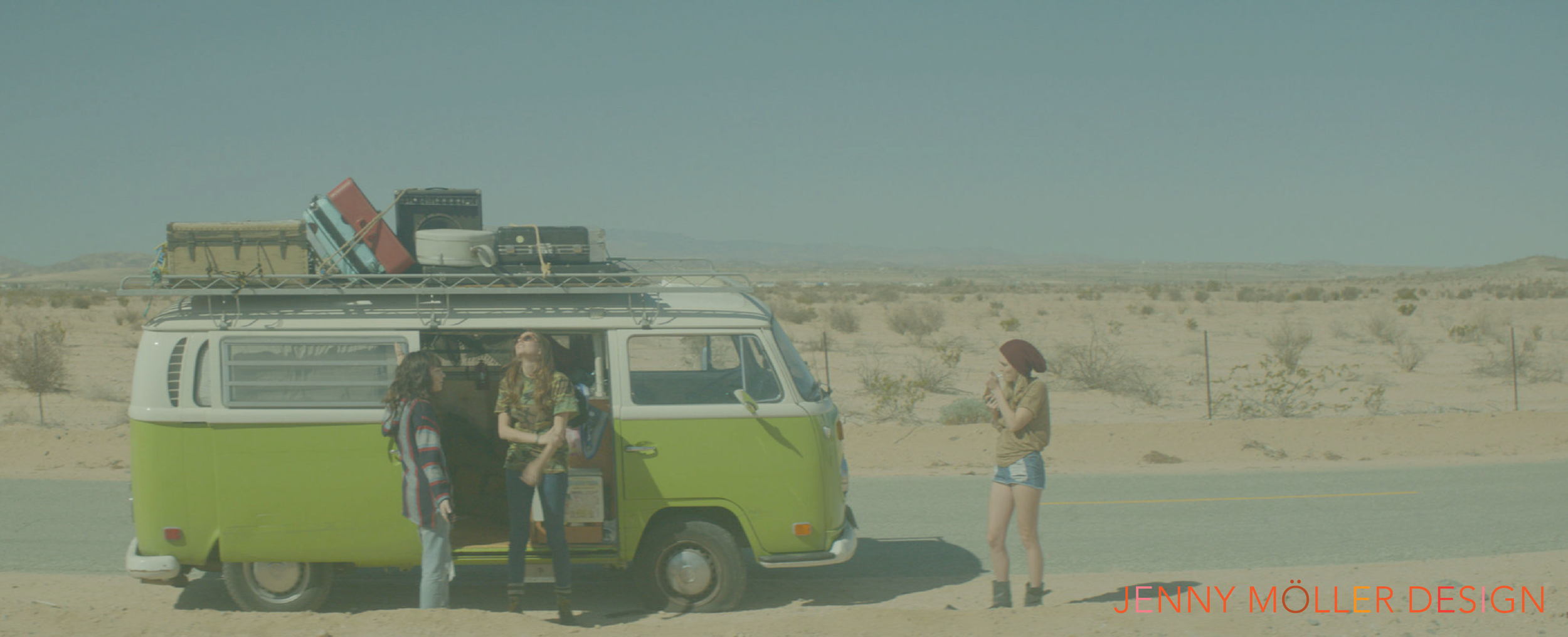Three women standing beside a green and white vintage camper van in a desert landscape, with luggage on the roof and one woman taking a photo with her phone.