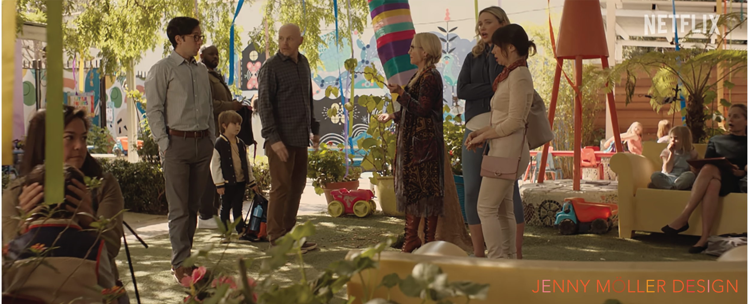 People gathered outdoors in a garden setting with colorful decorations, sitting on yellow couches and standing, engaged in conversation. There are children and adults, with trees and a mural in the background, and a logo for Netflix in the upper right corner.