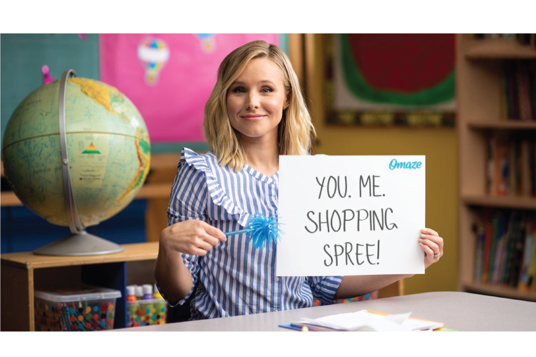 A woman sitting at a desk in a classroom, holding a sign that reads 'You. Me. Shopping. Spree!' with a small blue 'Omaze' logo. Behind her, there is a globe on a shelf and colorful classroom decorations.