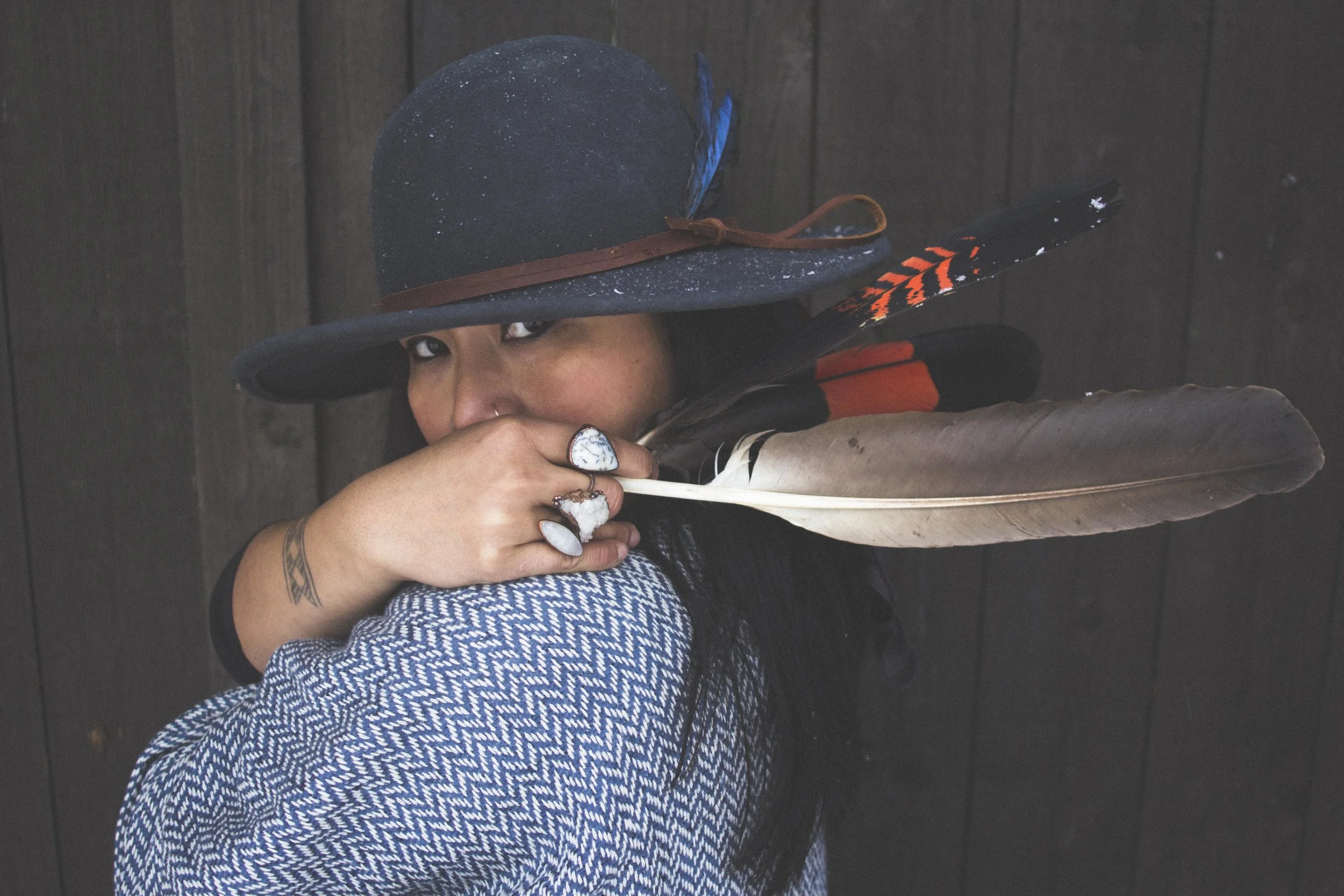 woman in blue and hat mysteriously looking out with hand covering mouth, feathers and crystal jewellery in hand