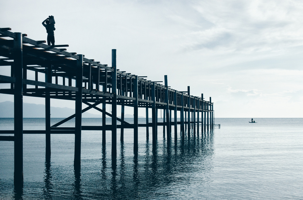 Early morning in Thailand with lone worker on pier and fisherman hoping to get an early catch. The air was warm, the water still and the clouds filtered the light to create a dream like scene.