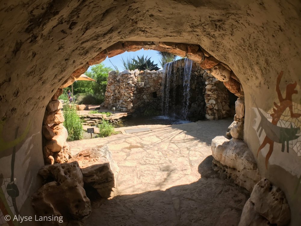  This was my favorite part of the garden, approaching as I did from the back. From that side, the tunnel’s destination and purpose is mysterious. The pictographs inside surprised and delighted me, and beyond—that waterfall! What had been hidden, unfo