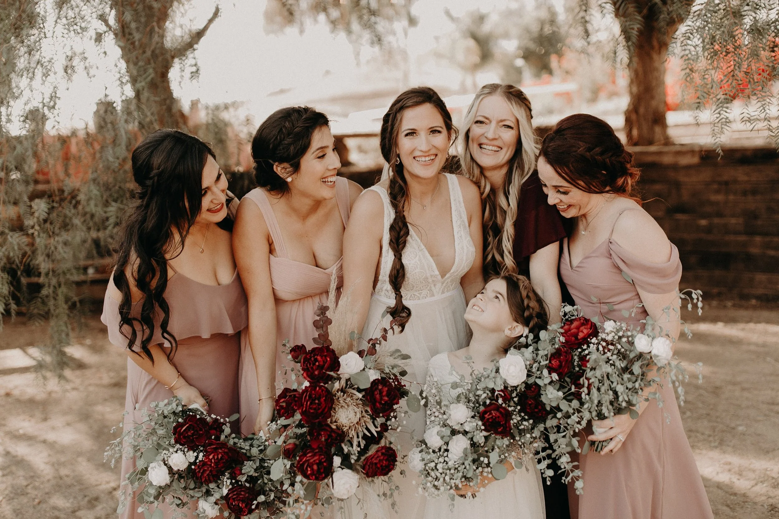 Bride smiling with her bridesmaids while holding burgundy, cream, and white wedding bouquets.