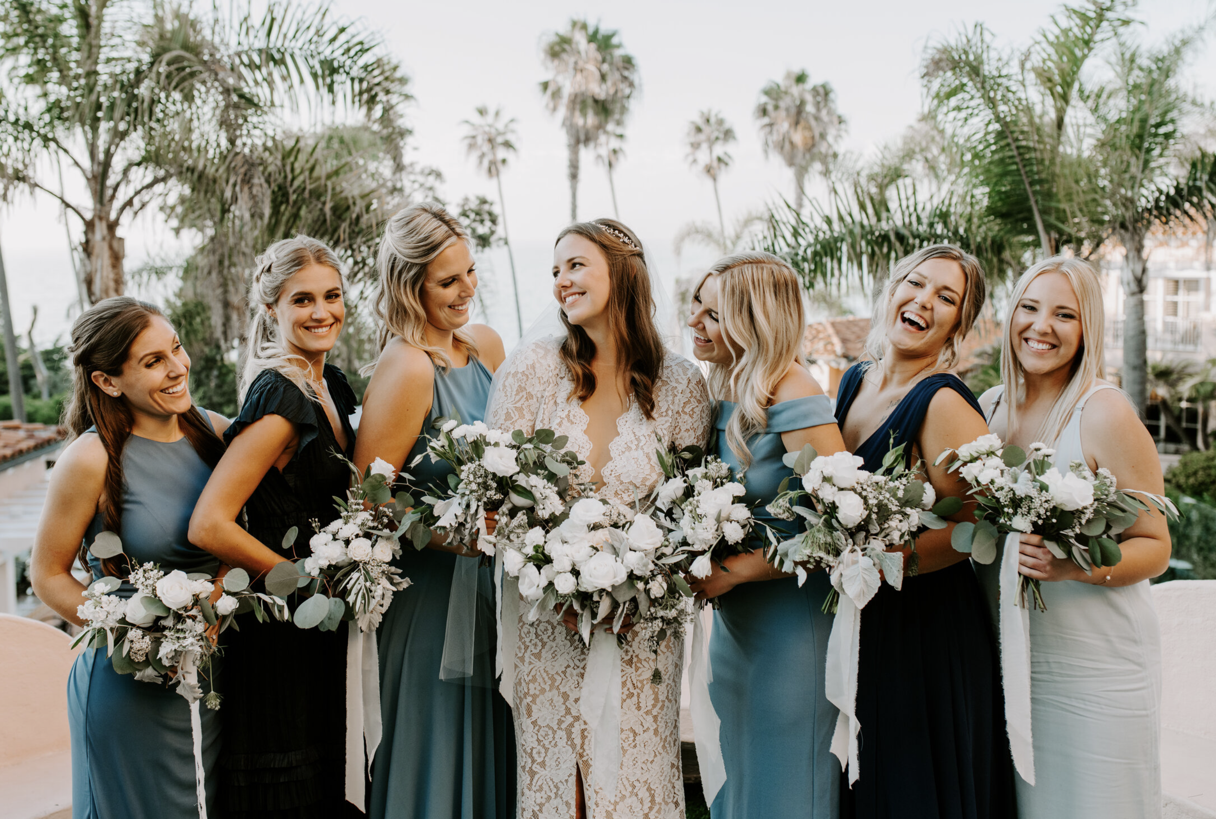 Bride laughing with her bridesmaids in teal and green dresses while holding white wedding bouquets outdoors.