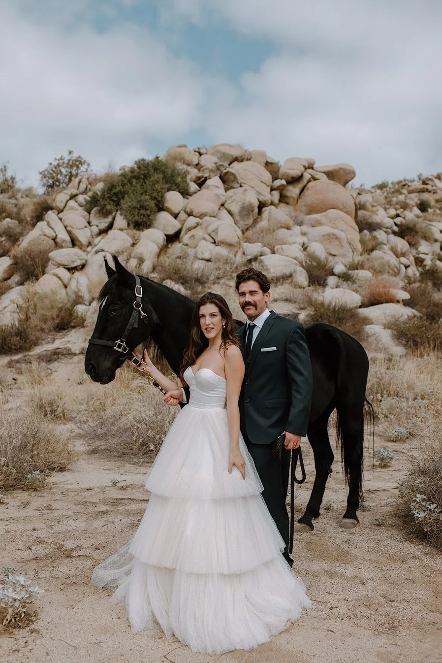Bride and groom standing beside a black horse in a rocky desert wedding setting.