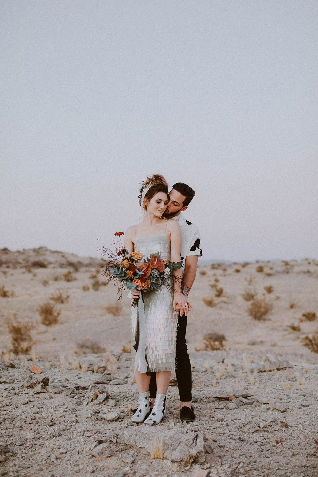 Bride and groom standing together in a desert setting with a rust-toned wedding bouquet.