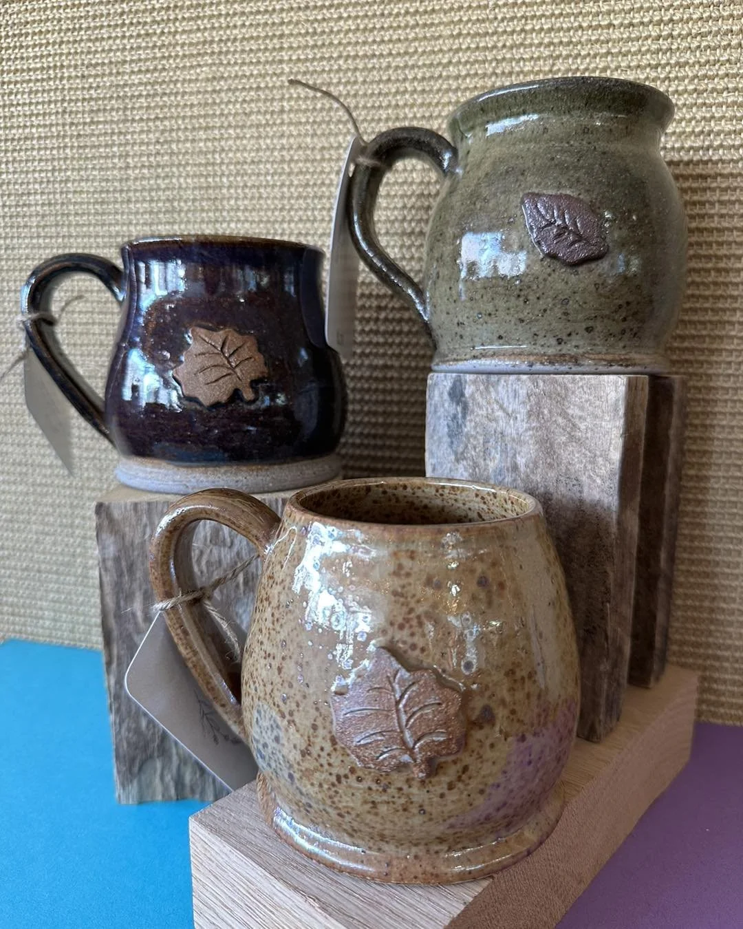 Three ceramic mugs with leaf designs, displayed on wooden blocks and a textured background.