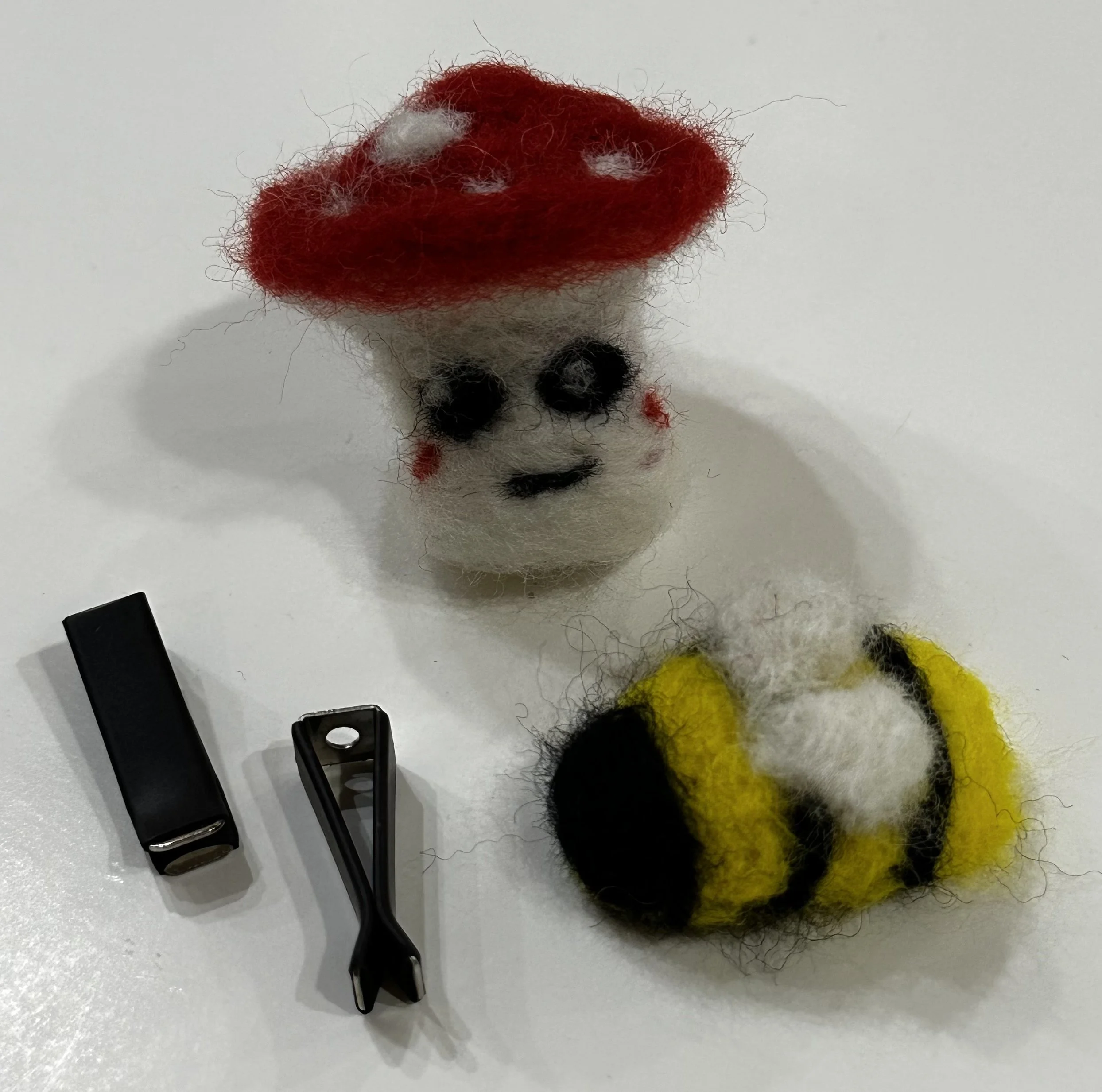A felted wool figure of a skeleton wearing a red hat with white spots, black eye sockets, red cheeks, and a yellow and black striped tail, along with a small black clip and a black box on a white surface.