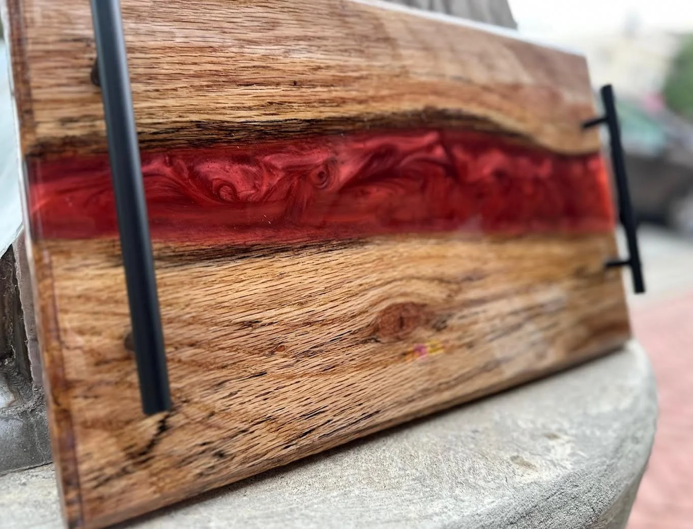 Close-up of a wooden cutting board with a red resin inlay in the center, resting on a concrete surface outside.