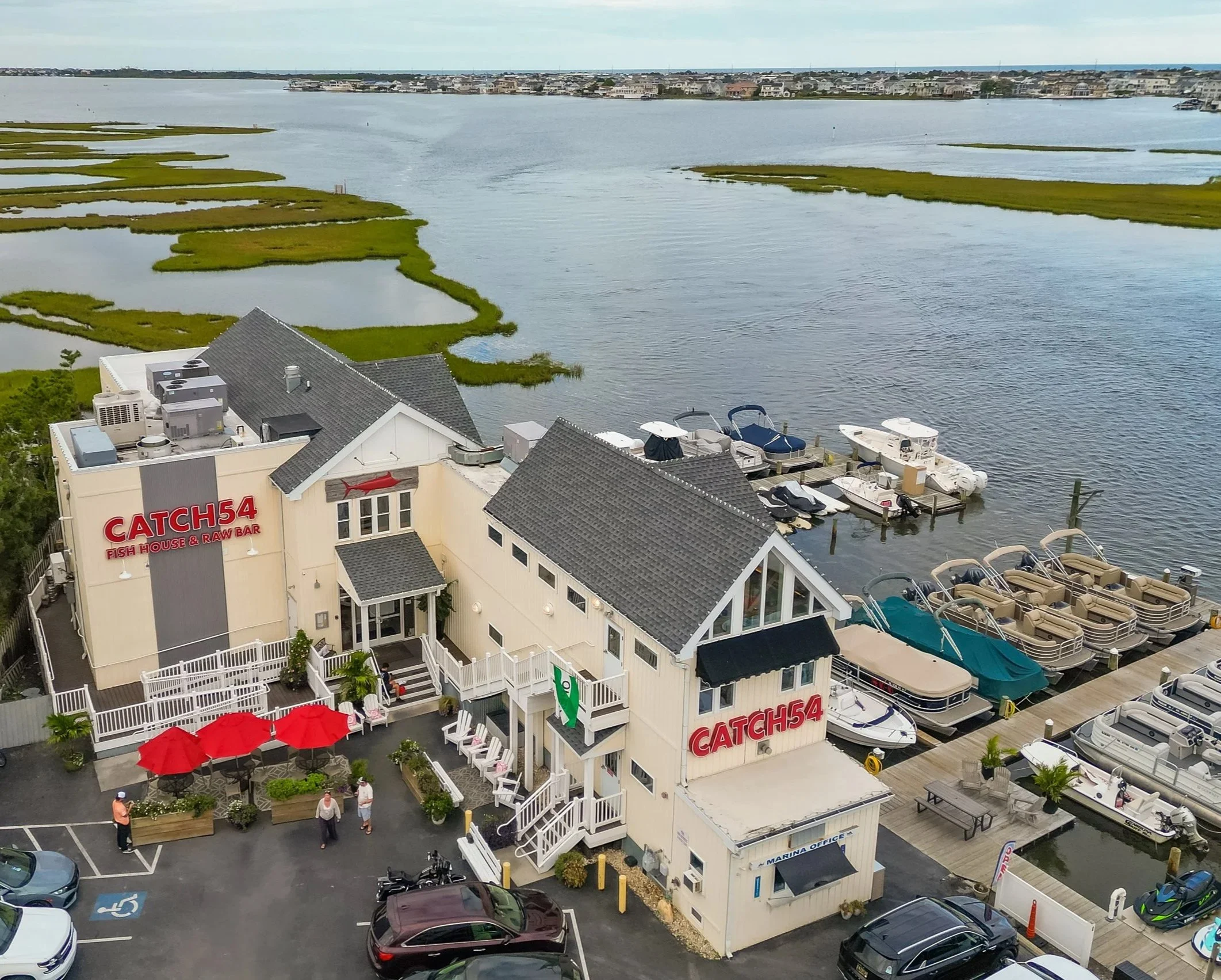 A marina with boats docked along the water, a restaurant called Catch 54 Fish House & Raw Bar with outdoor seating and red umbrellas, and a view of a body of water with grassy islands and a distant shoreline with houses.