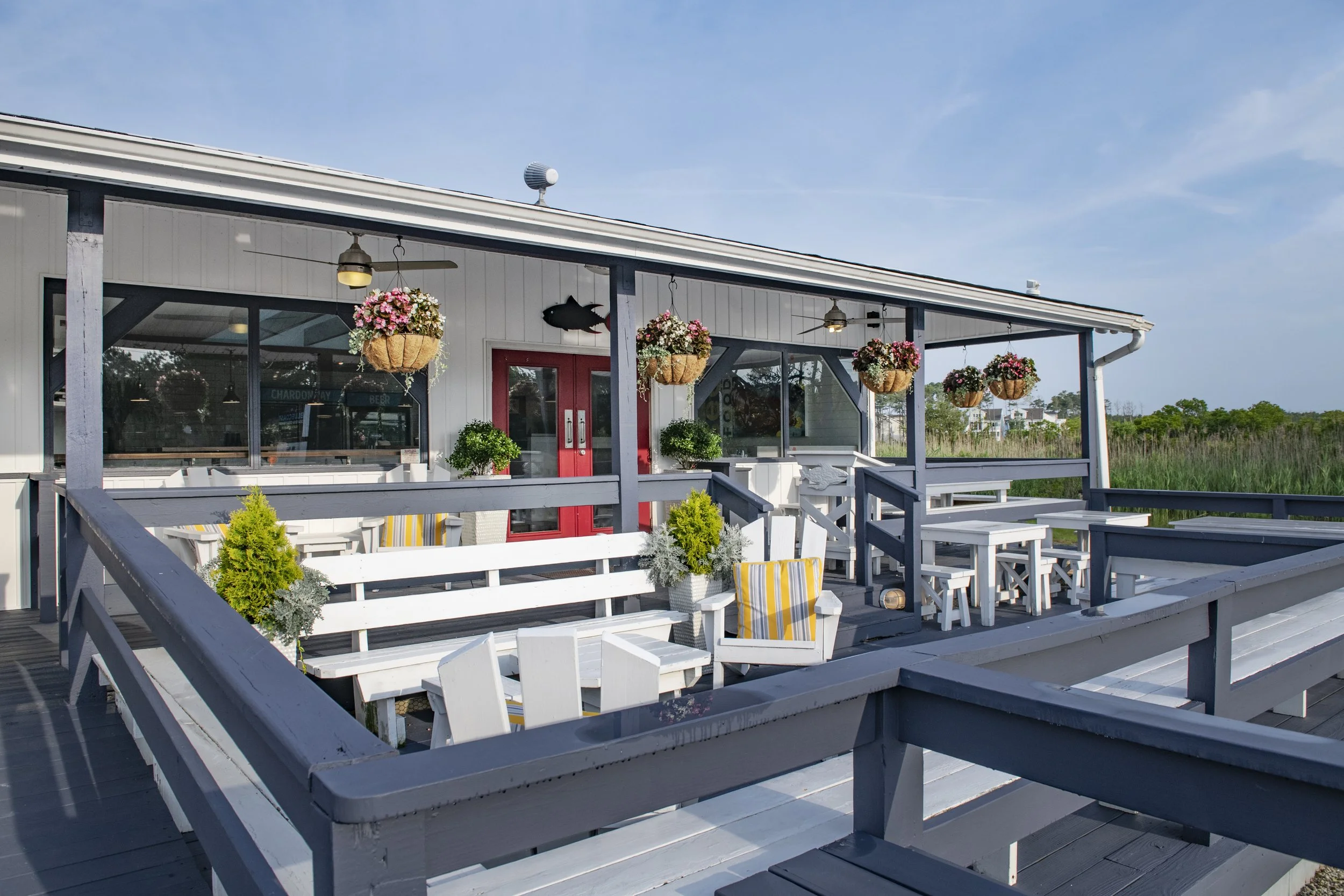 Front porch area of a seaside restaurant or cafe with hanging baskets of pink and white flowers, potted plants, white benches, and a red door.