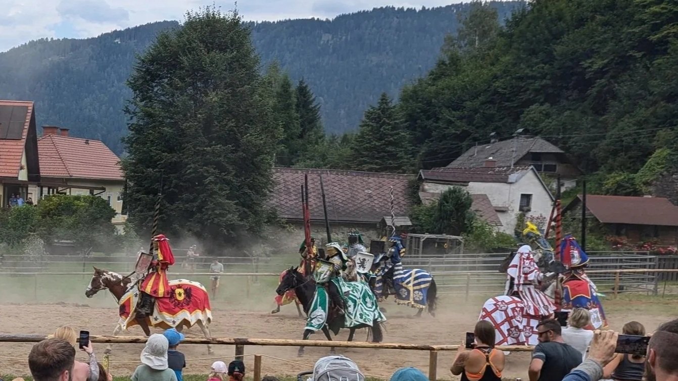 Ritter in mittelalterlicher Rüstung bei Turnier auf Pferden, im Hintergrund Häuser und Berglandschaft, Publikum beobachtet das Spektakel.